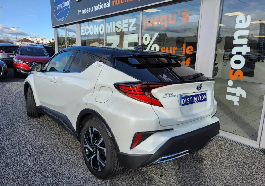 Toyota C-HR blanc nacré en 3/4 arrière droit, toit noir et jantes alliage 18 pouces visibles devant un showroom.
