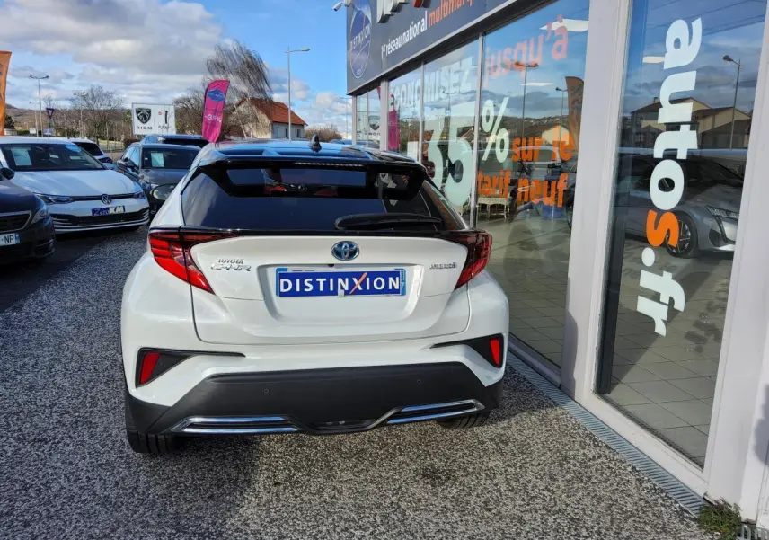 Vue arrière d'un Toyota C-HR blanc nacré avec ornements chromés, stationné devant une vitrine de concession multimarque.