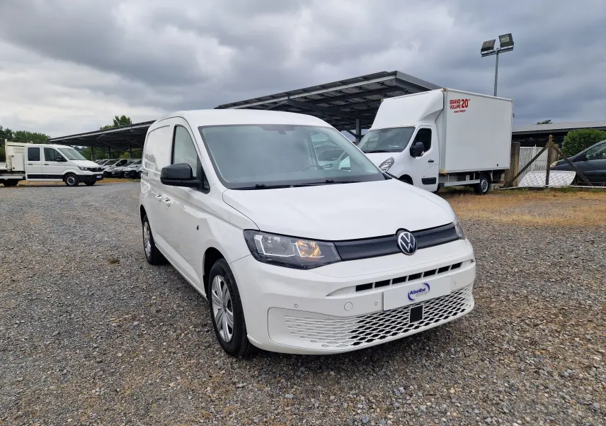 Volkswagen Caddy Cargo blanc vu en 3/4 avant droit sur un sol gravillonné avec plusieurs utilitaires en arrière-plan.