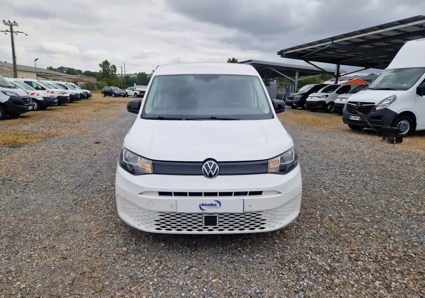 Vue frontale d'un Volkswagen Caddy Cargo blanc 2023 sur un parking avec plusieurs autres utilitaires autour.