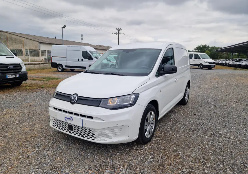 Volkswagen Caddy Cargo blanc vu en 3/4 avant droit sur un sol gravillonné avec plusieurs utilitaires en arrière-plan.