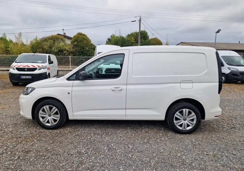 Vue de profil côté gauche d’un Volkswagen Caddy Cargo blanc 2023 sur un sol gravillonné, sans marquages visibles.