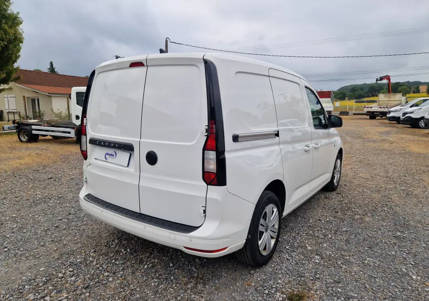 Vue 3/4 arrière droite d'un Volkswagen Caddy Cargo blanc 2023 sur un sol gravillonné, avec portes arrière fermées.