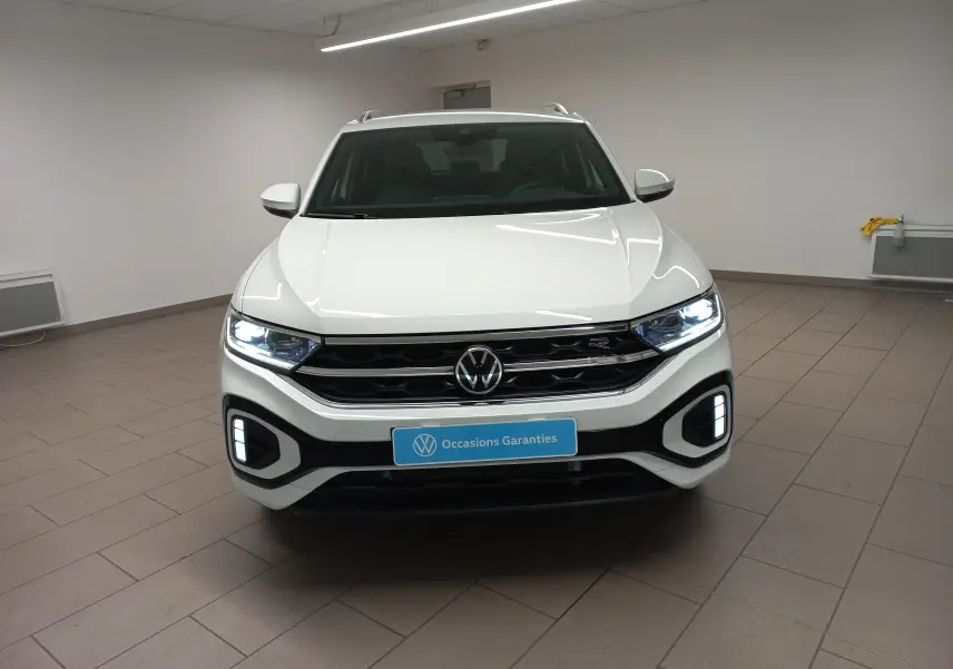 Vue frontale d'un Volkswagen T-Roc blanc pur 2025 avec feux LED allumés et calandre R-Line dans un showroom.