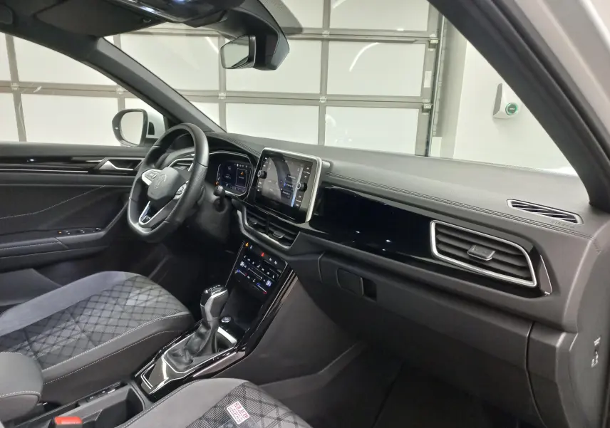 Vue intérieure côté conducteur du Volkswagen T-Roc 1.5 TSI blanc, avec tableau de bord moderne et sièges tissu à motifs.