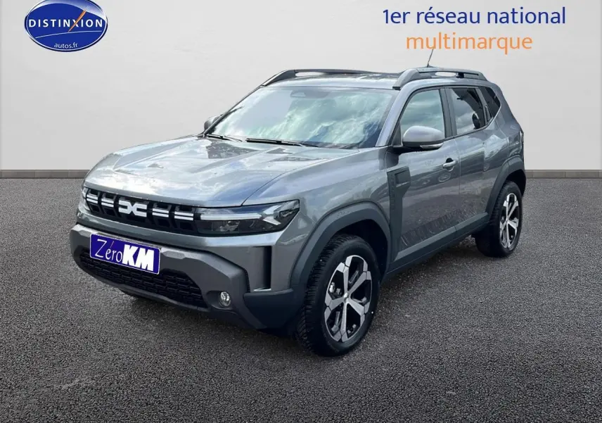 Vue 3/4 avant du Dacia Duster gris schiste 2025 avec jantes alu et barres de toit visibles sur sol bitumé.