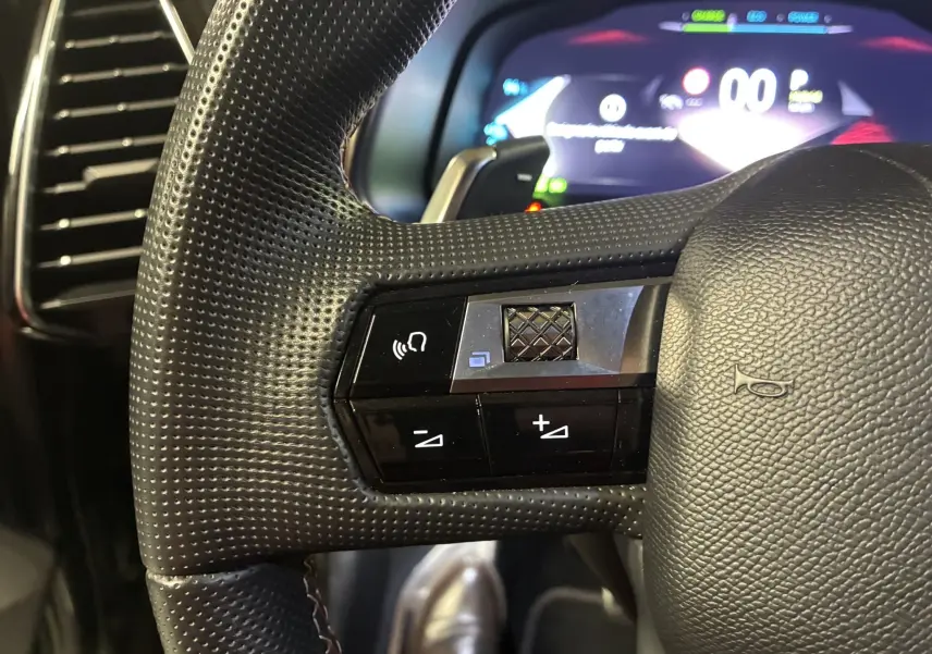 Gros plan sur la partie gauche du volant noir perforé du DS7 E-TENSE 2024 avec commandes intégrées et tableau de bord numérique flou.