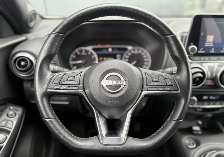 Vue rapprochée du volant en cuir noir du Nissan Juke blanc perle 2024, avec commandes intégrées et tableau de bord flou.
