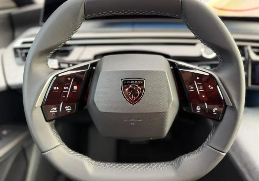 Gros plan sur le volant noir cuir du Peugeot 3008 Hybrid 2025 avec commandes tactiles et logo lion central.