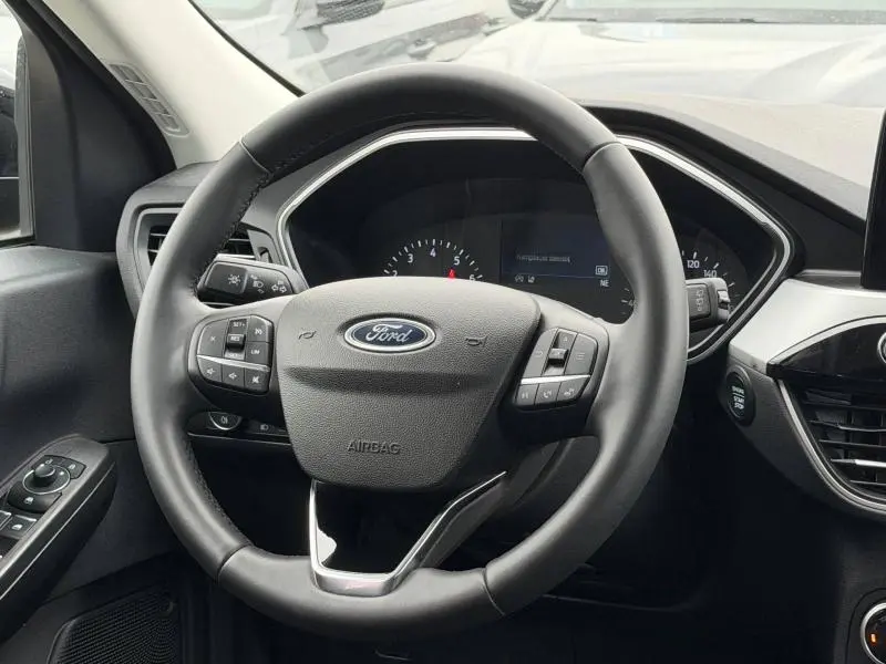 Vue intérieure centrée sur le volant noir du Ford Kuga 2024, avec commandes intégrées et tableau de bord partiellement visible.