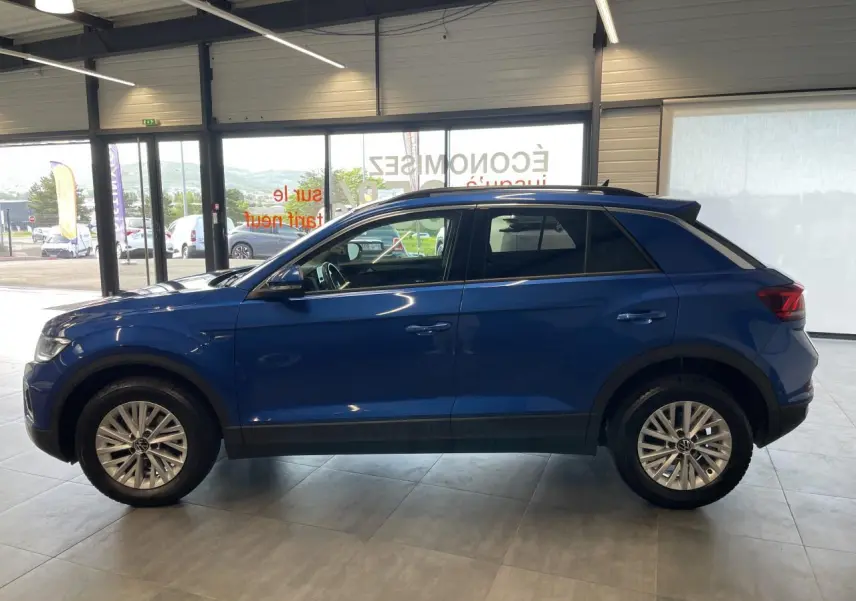Volkswagen T-Roc bleu en vue de profil côté gauche, avec jantes alliage et vitres teintées en showroom.
