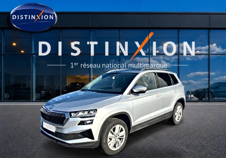 Vue 3/4 avant droite d'un Skoda Karoq gris 2025 devant une vitrine Distinxion, avec calandre noire et jantes alu.