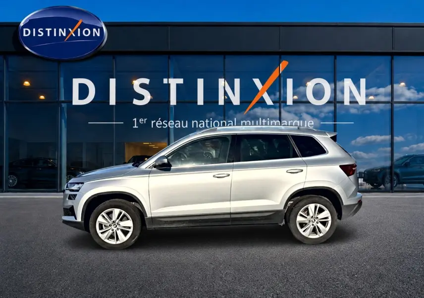 Vue de profil côté gauche d'un Skoda Karoq gris 2025 garé devant une vitrine avec logo Distinxion.