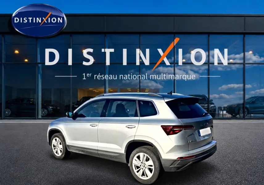 Vue 3/4 arrière droite d’un Skoda Karoq gris 2025 garé devant une vitrine avec logo Distinxion.