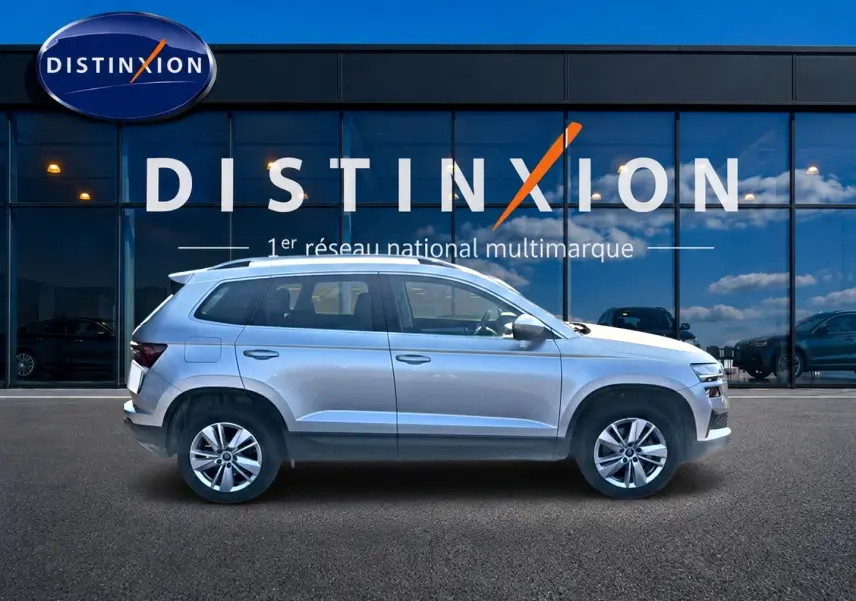 Profil droit d'un Skoda Karoq gris 2025 garé devant une vitrine avec logo Distinxion visible en arrière-plan.