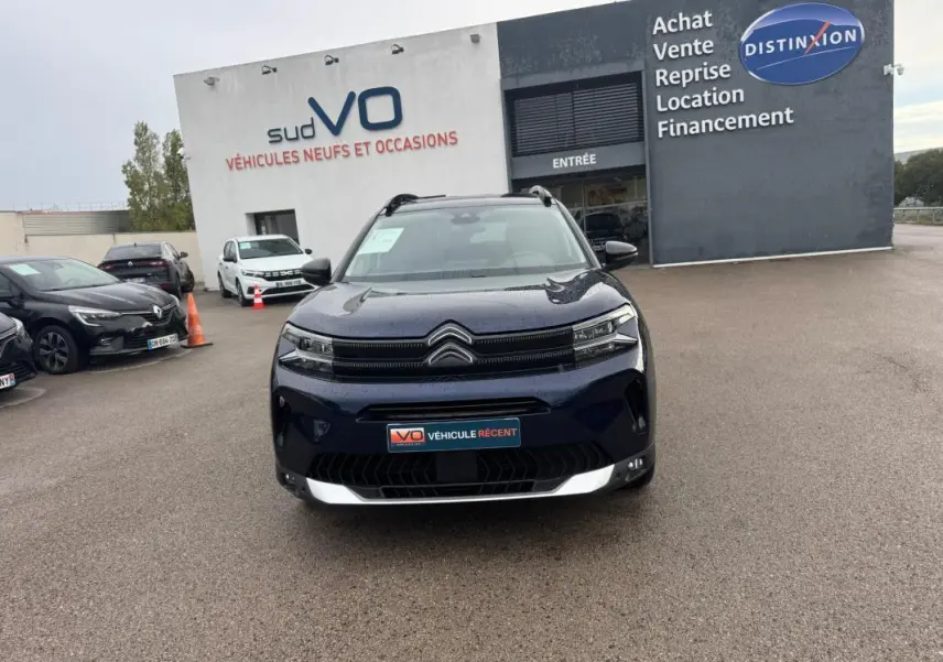 Citroën C5 Aircross hybride bleu foncé vue de face, avec barres de toit et feux allumés sur parking de concession.