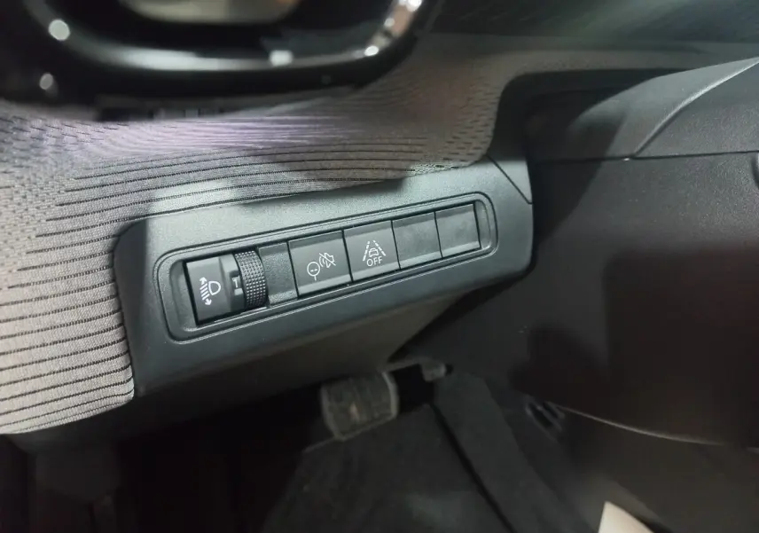 Gros plan sur les commandes de réglage des phares et aides à la conduite côté gauche du tableau de bord de la Citroën C3 gris foncé.