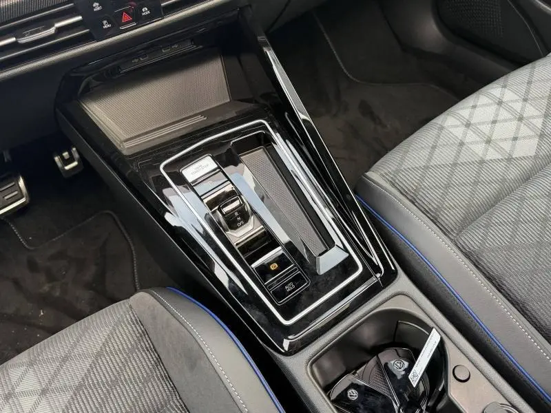 Vue rapprochée de la console centrale noire brillante avec levier de vitesse automatique et sièges gris à surpiqûres bleues de la Volkswagen Golf 2025.