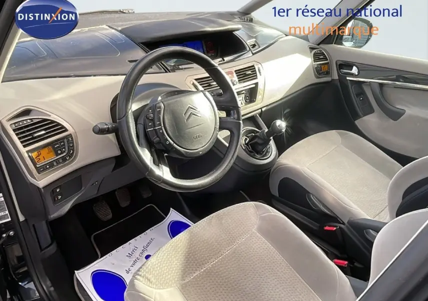 Vue intérieure côté conducteur de la Citroën C4 Picasso 2008, volant cuir noir et sièges tissu beige clair.