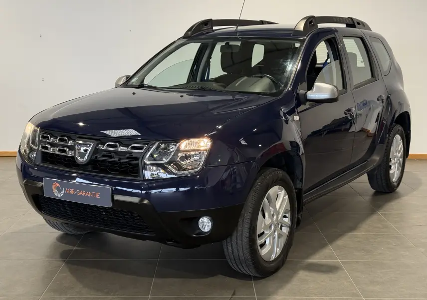 Dacia Duster bleu en 3/4 avant droit, avec calandre chromée et phares allumés dans un intérieur neutre.