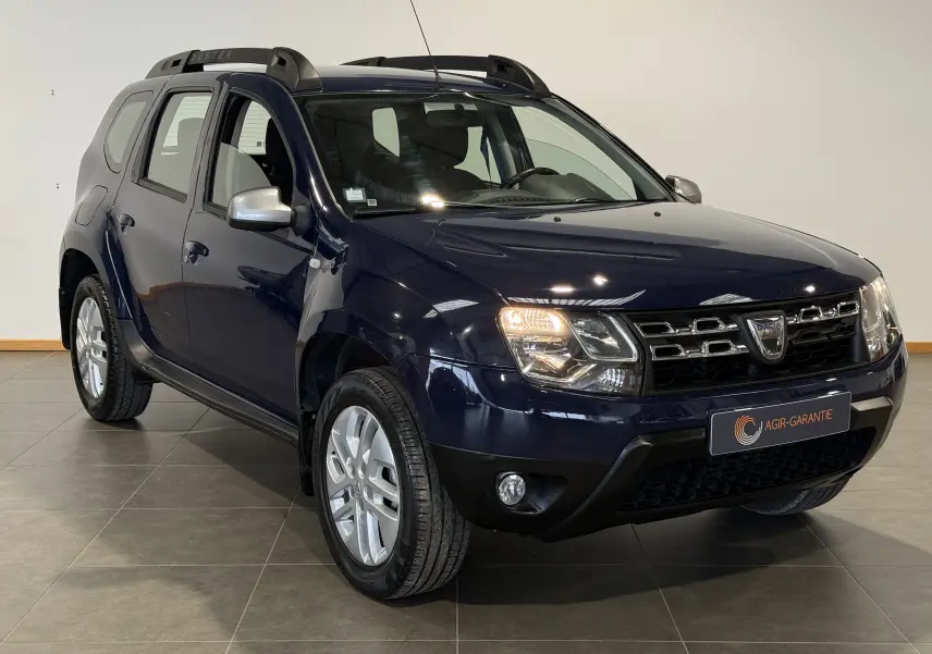 Dacia Duster bleu en 3/4 avant droit, avec calandre chromée et barres de toit noires visibles en intérieur showroom.