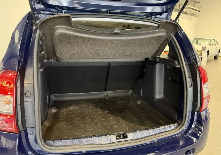 Coffre ouvert vu de l'arrière d'un Dacia Duster bleu 2015, montrant l'espace de chargement vide et la tablette arrière relevée.