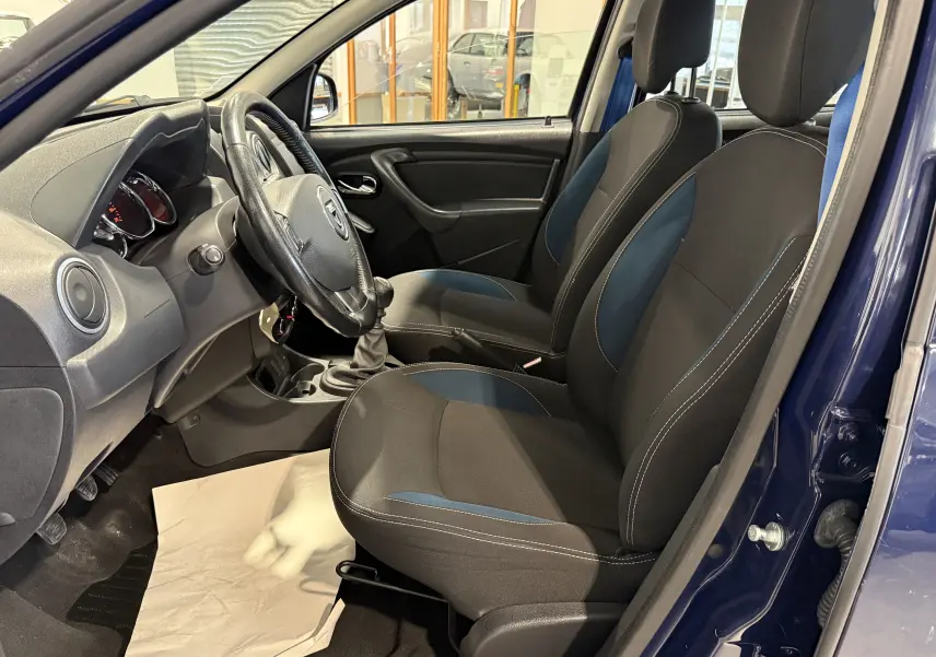 Vue intérieure côté conducteur du Dacia Duster bleu 2015, montrant les sièges tissu noirs avec inserts bleus et volant cuir.