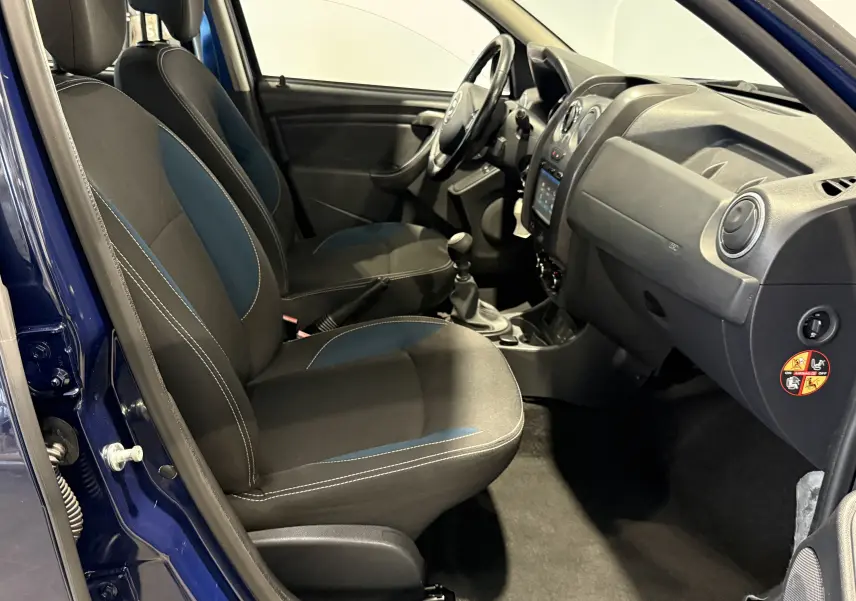 Vue intérieure côté conducteur du Dacia Duster bleu 2015, montrant sièges avant, tableau de bord et levier de vitesse manuel.