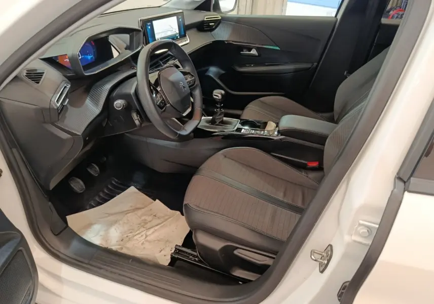 Vue intérieure côté conducteur de la Peugeot 208 blanche, montrant le tableau de bord numérique et la console centrale avec boîte manuelle.