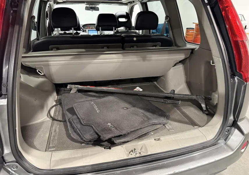 Vue arrière du coffre ouvert d’un Nissan X-Trail Columbia gris 2006 avec tapis de sol et cache-bagages visibles.