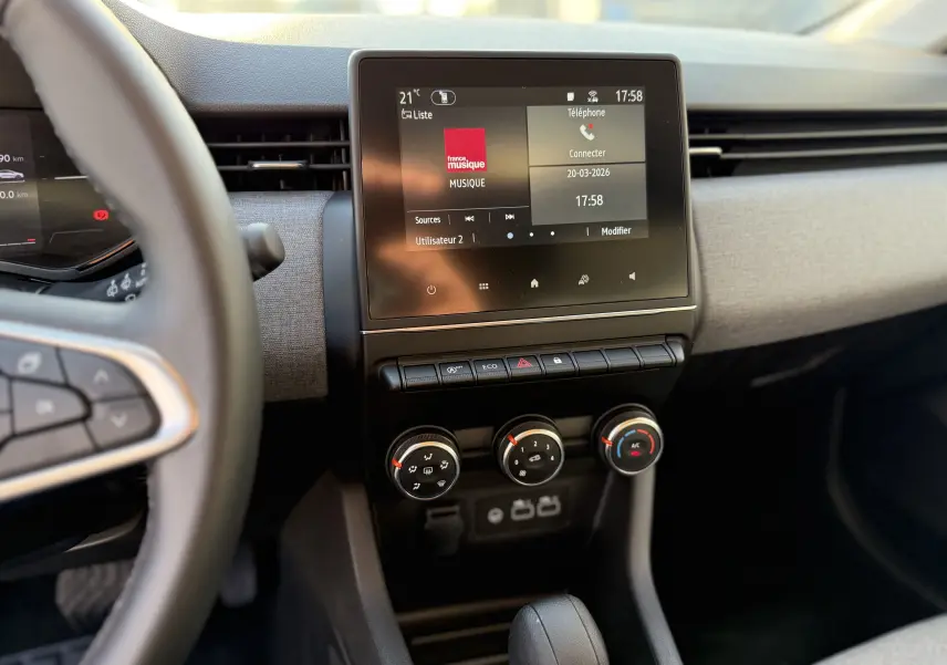 Vue intérieure centrée sur l’écran tactile et la console centrale de la Renault Clio V gris schiste, avec commandes de climatisation visibles.