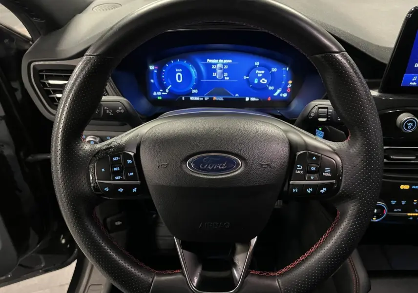 Vue rapprochée du volant noir perforé avec surpiqûres rouges et tableau de bord numérique bleu du Ford Kuga 2022 ST-Line.