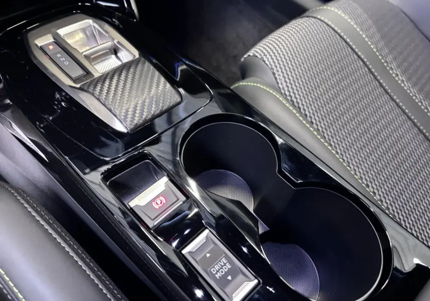Gros plan sur la console centrale noire brillante avec sélecteur de boîte auto et bouton Drive Mode, siège tissu gris avec surpiqûres vertes.