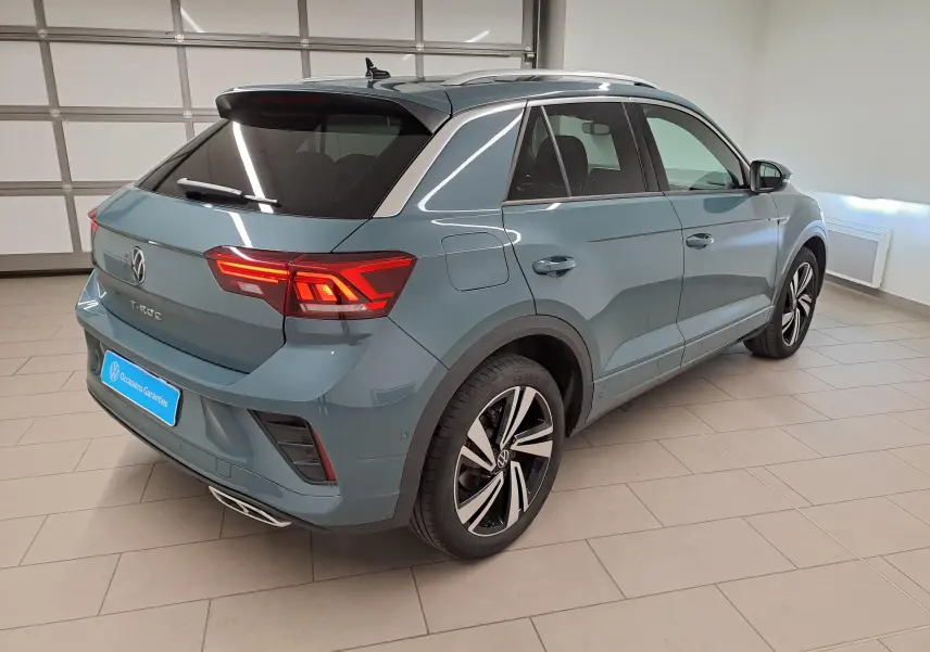 Vue 3/4 arrière droite d’un Volkswagen T-Roc bleu pétrole 2025 avec jantes alliage et feux arrière allumés dans un garage.