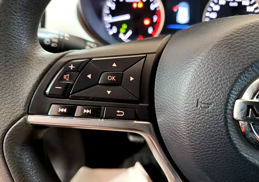 Gros plan sur le volant multifonctions noir de la Nissan Micra IG-T 100 Business Edition avec commandes audio et tableau de bord flou.