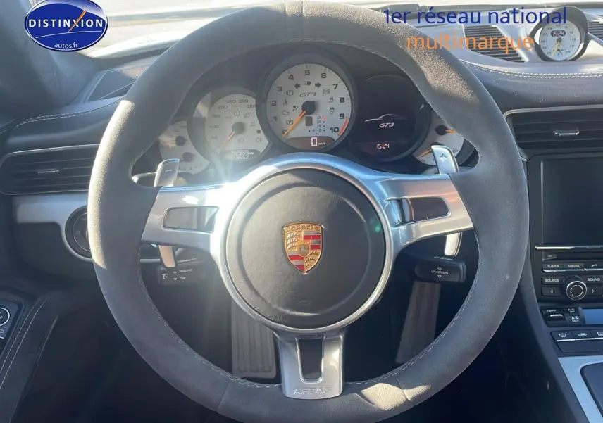 Vue intérieure centrée sur le volant en alcantara gris d’une Porsche 911 GT3 blanc, avec compte-tours et tableau de bord visibles.