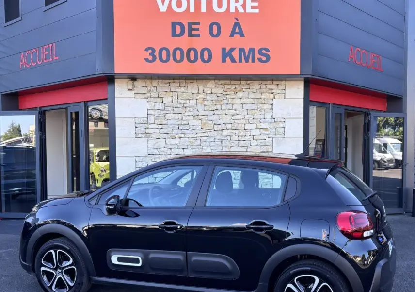 Profil latéral droit d'une Citroën C3 noire 2024 avec protections latérales caractéristiques, stationnée devant un bâtiment commercial.