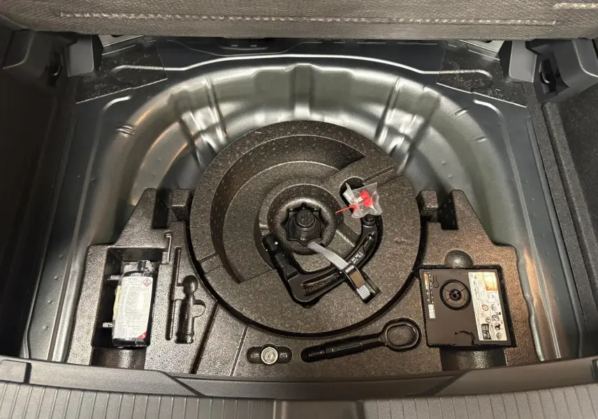 Vue du coffre ouvert du Volkswagen T-Roc 2025 gris Indium, montrant le rangement sous le plancher avec dispositif d'attelage amovible.