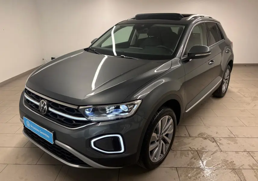 Volkswagen T-Roc 2025 gris Indium en 3/4 avant droit avec feux allumés et attelage amovible visible.