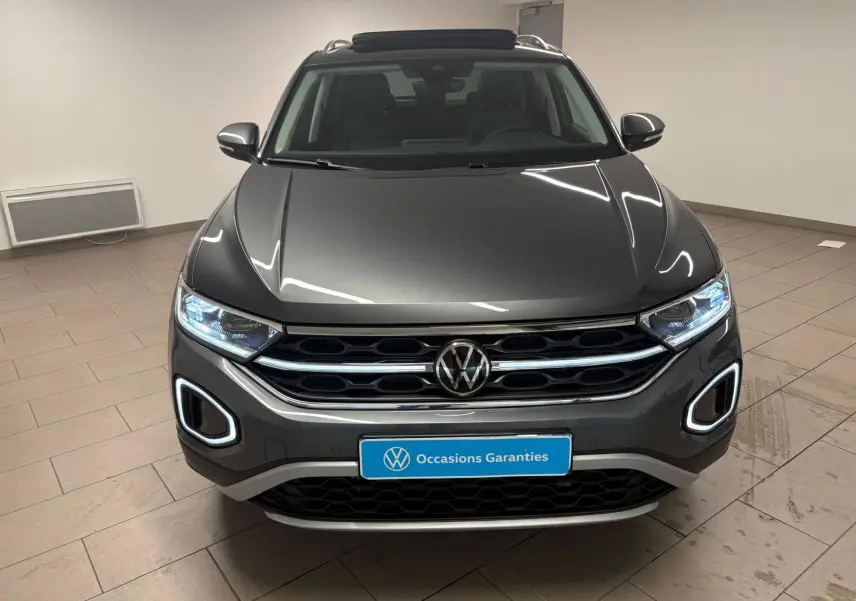 Vue avant d'un Volkswagen T-Roc 2025 gris Indium avec feux allumés et attelage amovible visible en intérieur.