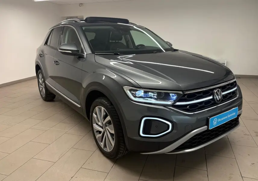 Volkswagen T-Roc 2025 gris Indium en 3/4 avant droit, avec phares LED et attelage amovible visible.
