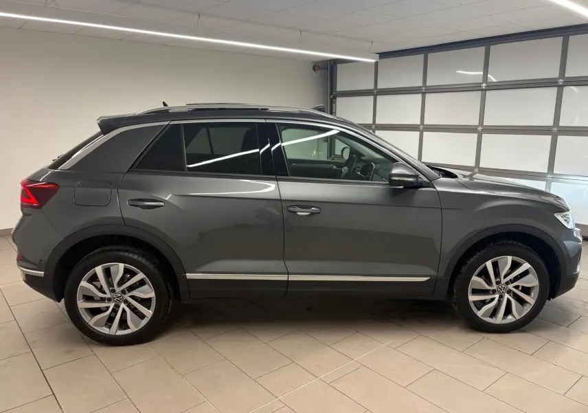Volkswagen T-Roc 2025 gris Indium vu de profil côté gauche, avec attelage amovible visible à l'arrière.