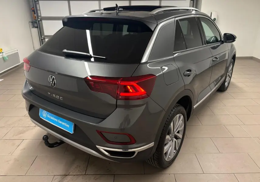 Vue 3/4 arrière droite du Volkswagen T-Roc gris Indium avec attelage amovible et feux arrière allumés en intérieur.