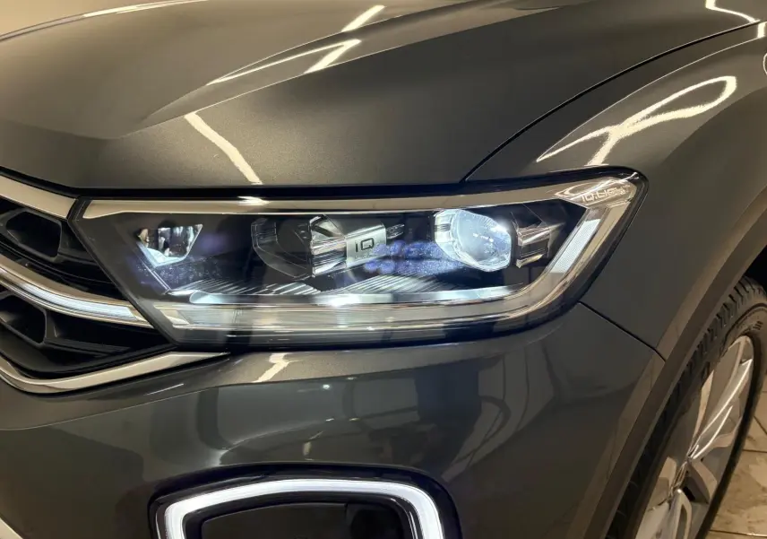 Gros plan sur le phare avant droit du Volkswagen T-Roc 2025 gris Indium avec peinture métallisée et jante visible.