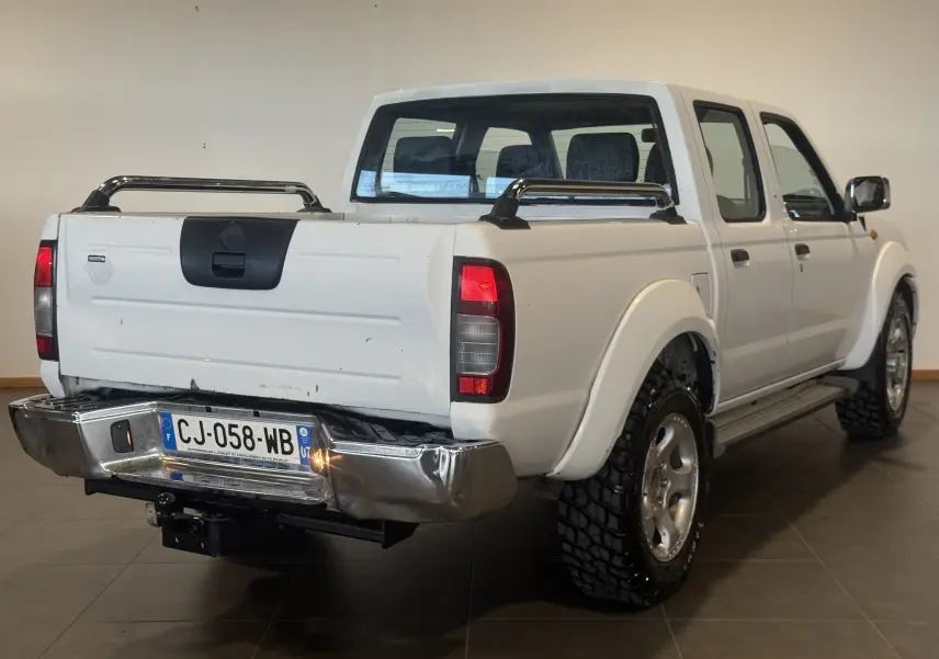 Vue 3/4 arrière droite d’un Nissan Navara NP300 blanc de 2002 avec benne double cabine et pare-chocs arrière chromé abîmé.