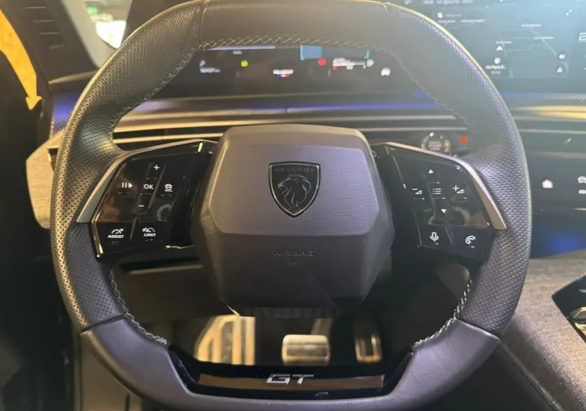 Volant cuir perforé du Peugeot 5008 1.2 HYBRID 136 GT avec logo lion et inscription GT, tableau de bord numérique en arrière-plan.