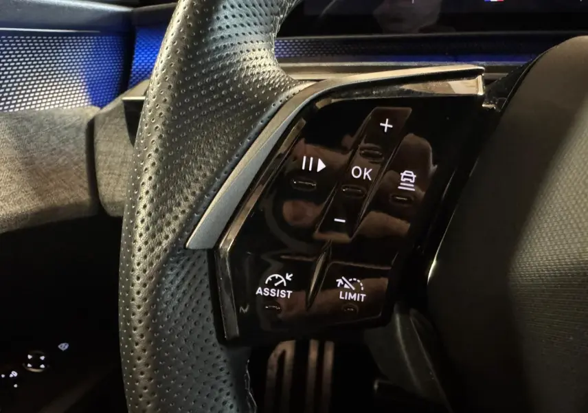 Gros plan sur les commandes au volant côté gauche du Peugeot 5008 gris 1.2 HYBRID 136 GT, affichage digital en arrière-plan.