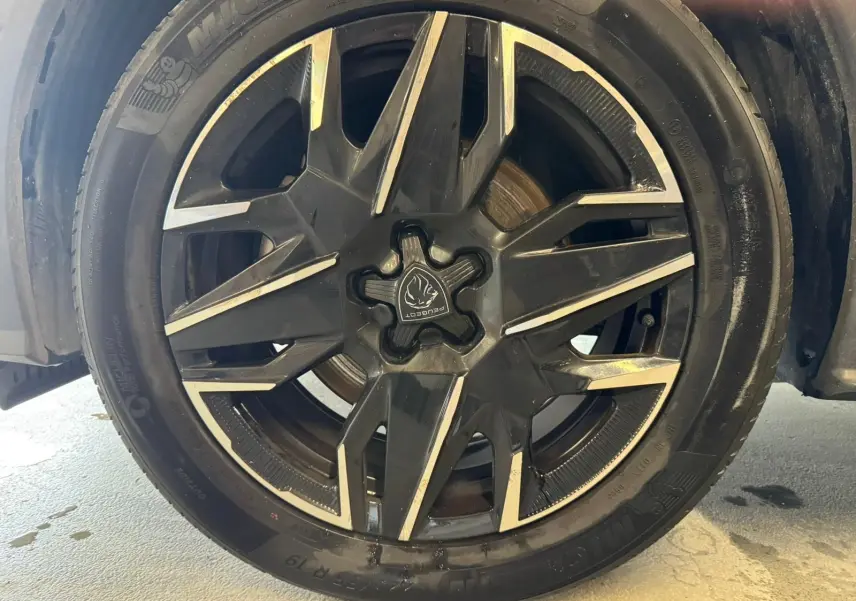 Gros plan sur la jante noire et argentée du Peugeot 5008 1.2 HYBRID 136 GT gris, avec logo Peugeot central visible.