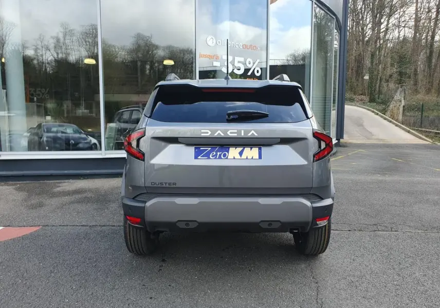 Vue arrière d'un Dacia Duster gris schiste 2026 avec feux LED et plaque d'immatriculation Zero KM devant un bâtiment vitré.