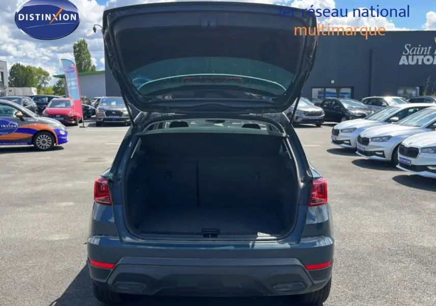Vue arrière d'un SEAT Arona bleu Fjord avec coffre ouvert, toit blanc visible, sur un parking extérieur ensoleillé.